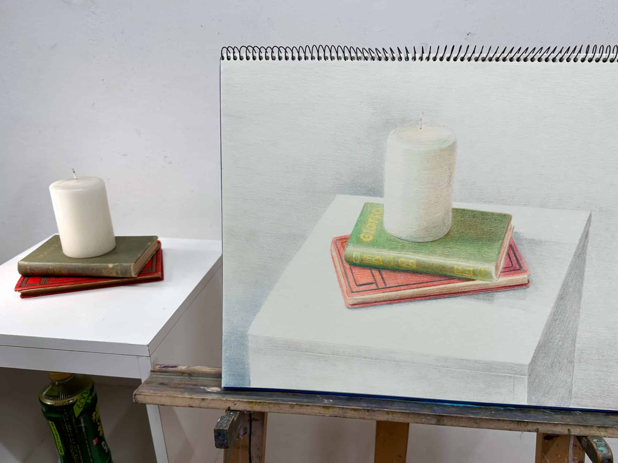 Colored pencil still life drawing by Alessandro Cuzzocrea. The drawing, displayed on a sketchpad, shows a realistic depiction of a white candle resting on top of two books with green and red covers. The artwork is placed on an easel, and the actual setup of the candle and books is visible beside it, allowing for a comparison between the drawn and real objects