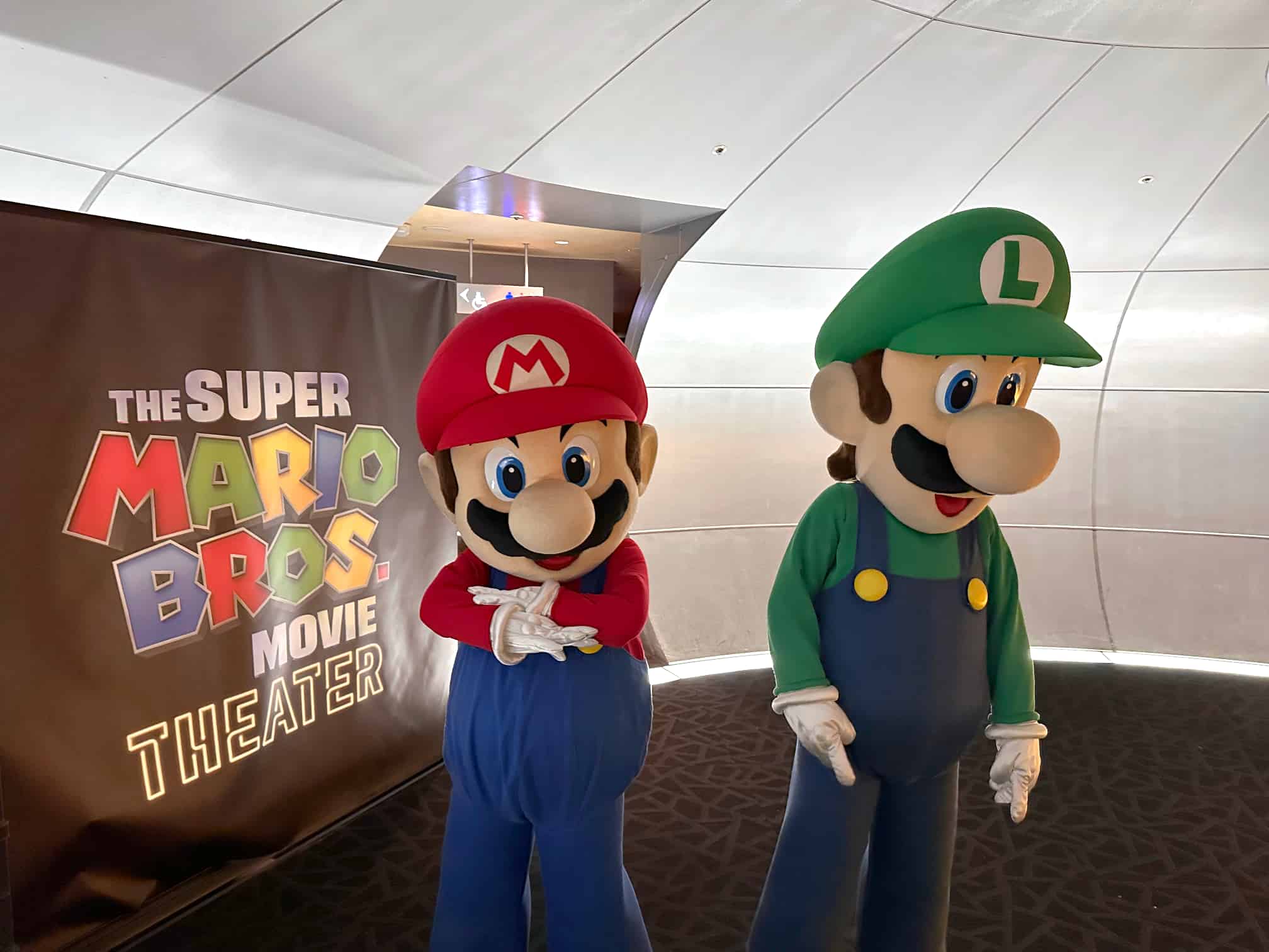 Mario and Luigi from the Super Mario franchise standing in a theater lobby, with a backdrop promoting The Super Mario Bros. Movie. Mario is on the left, wearing his signature red hat and blue overalls, while Luigi is on the right, in green and blue attire. They appear to be posing for a photograph